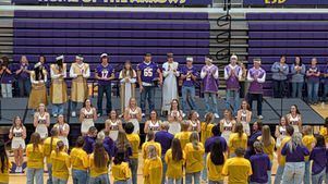 Cheerleaders in white uniforms, students in purple jerseys, and gold robes stand on bleachers and stage, clapping in a gymnasium.