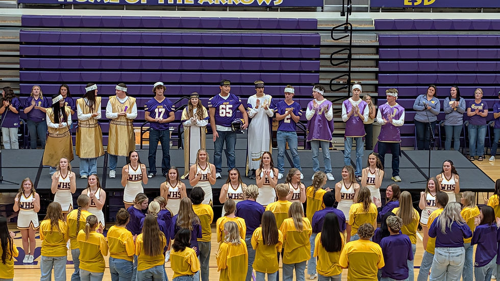 Cheerleaders in white uniforms, students in purple jerseys, and gold robes stand on bleachers and stage, clapping in a gymnasium.