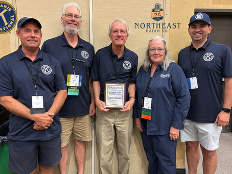 Five Kiwanis members pose in a hallway; one holds a Kiwanis award plaque, with Northeast Radio SD and Kiwanis logos behind.