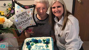 Smiling caregiver poses with an elderly woman beside a 100th birthday cake and balloon reading happy 100th birthday.