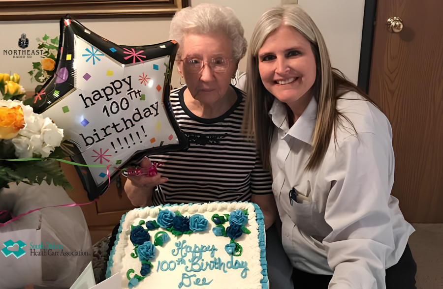 Smiling caregiver poses with an elderly woman beside a 100th birthday cake and balloon reading happy 100th birthday.