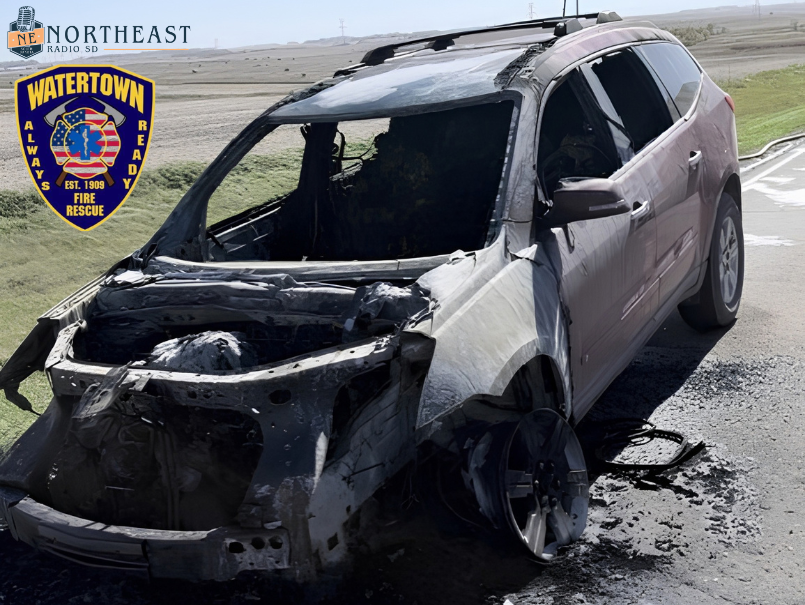 Burned-out SUV with shattered windows and missing wheel on roadside; Northeast Radio SD and Watertown Fire Rescue logos at left.