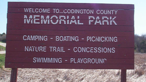 Red wooden sign reads: "Welcome to Codington County Memorial Park." Lists activities: camping, boating, picnicking, nature trail, concessions, swimming, playground. Set against a grassy field.