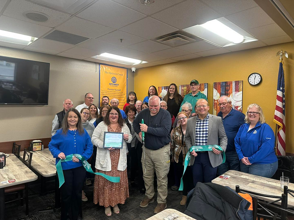 Group of people in a room with yellow walls, smiling and holding a ribbon for a ceremony. "The Optimist Creed" poster and U.S. flag visible.