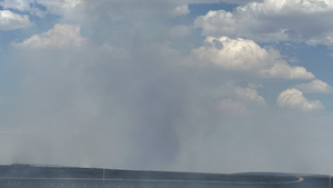 Cloudy sky with smoke rising from a charred landscape below. Sparse fencing and a distant horizon suggest a vast, open area.