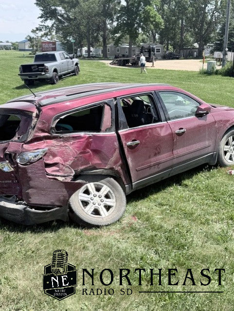 Damaged maroon SUV in a grassy yard after a crash, with a tow truck and people in the background; Northeast Radio SD logo visible.
