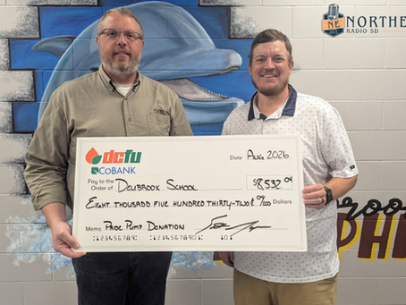 Two men hold a large check for Deubrook School, memo Pride Pump Donation, in front of a dolphin mural and Northeast Radio SD logo.