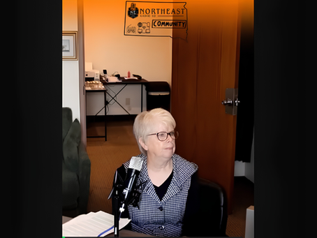 Older woman with glasses sits at a microphone in a doorway, with a Northeast Community Radio SD sign overhead.