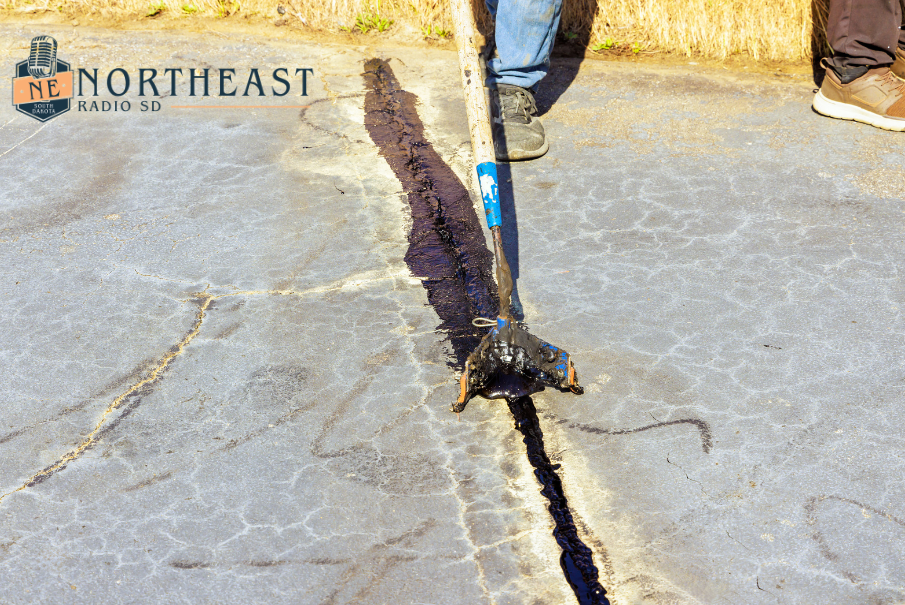 Person uses a long tool to seal a cracked pavement with black tar; Northeast Radio SD logo at top left.