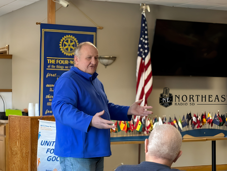Man in blue jacket speaks at Rotary meeting with gestures, in front of flags and screen labeled NORTHEAST RADIO SD.