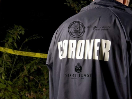 Back of a coroner in a dark jacket marked CORONER, standing by yellow police tape in a dark wooded area at night.