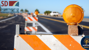 Roadwork zone with orange-striped barriers and flashing lights. "SD Department of Transportation" logo in corner. Sunny day, clear road.