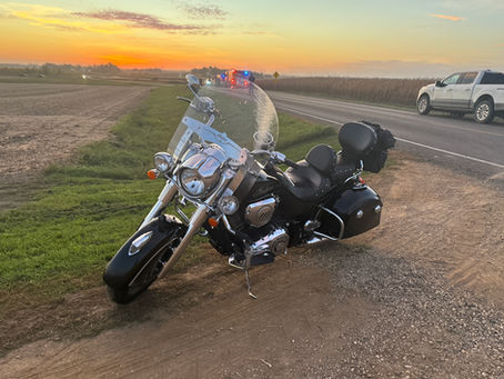 Black Indian motorcycle parked on a rural roadside at sunset, with flashing emergency vehicles in the distance and a calm, open field backdrop.