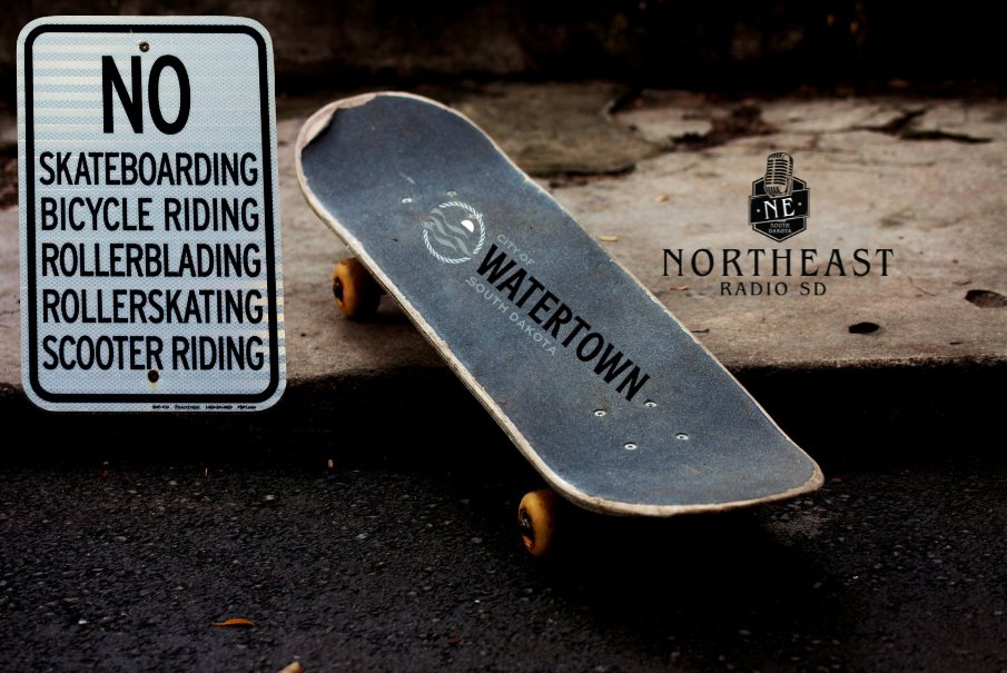 Skateboard leans on a curb beside a sign reading NO SKATEBOARDING, BICYCLE RIDING, ROLLERBLADING, ROLLERSKATING, SCOOTER RIDING.