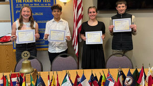 Four people smile holding "Rotary Honor Roll" certificates. Background shows a "Four-Way Test" banner, flags, and an American flag.