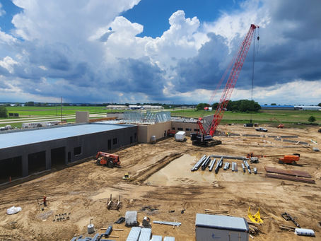 Large building under construction with a red crane and tractors on a muddy site beneath dramatic cloudy skies.