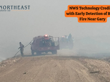 Firefighters use hoses to combat a brush fire in a smoky field. Red fire truck present. Text: NWS Technology Credited with Early Detection.