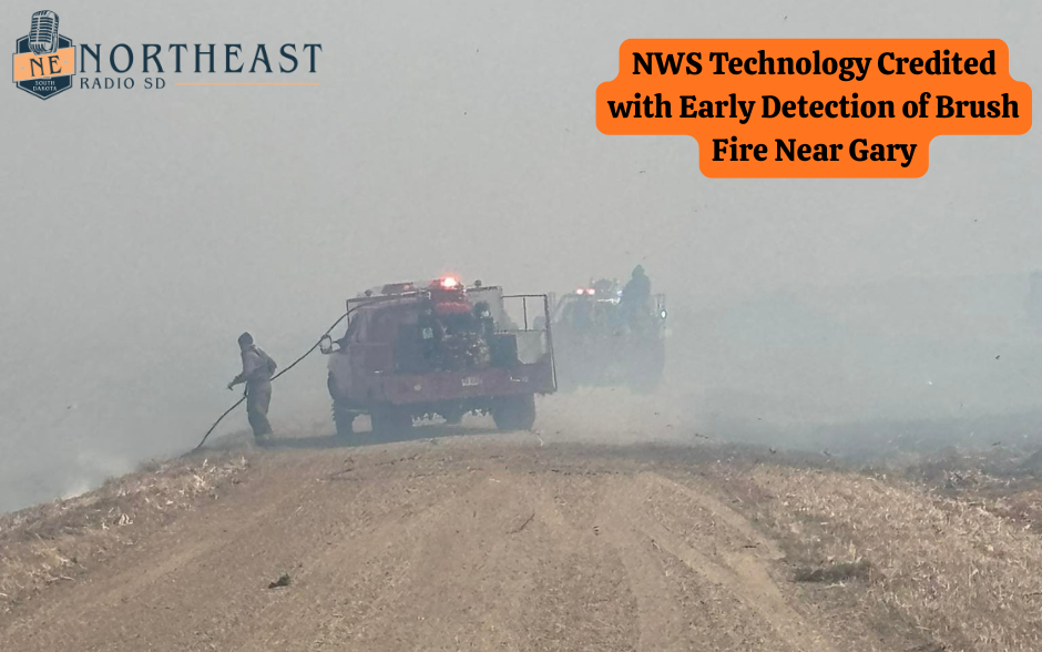 Firefighters use hoses to combat a brush fire in a smoky field. Red fire truck present. Text: NWS Technology Credited with Early Detection.