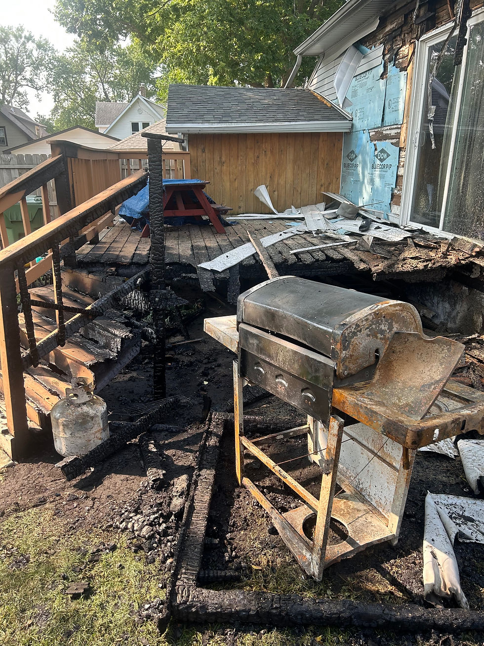 Burned backyard deck with a grill and propane tank, charred stairs, and damaged house siding after a fire.