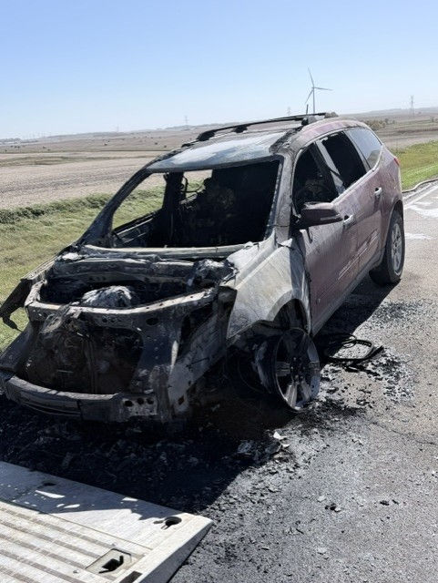 Burned SUV wreck on a rural roadside, front destroyed and wheels melted, with fields and a wind turbine in the background.