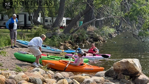 Kayakers launch colorful kayaks from a rocky riverbank under trees; Northeast Radio logo text appears at top left.