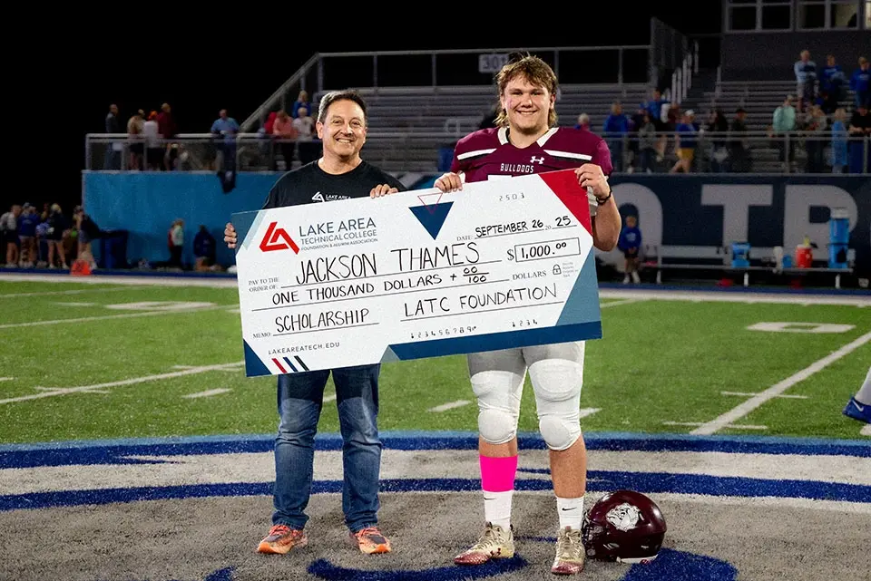 Two people on a football field holding a large scholarship check for $1,000 from Lake Area Technical College. Stadium seating in the background.