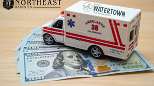 Toy ambulance labeled City of Watertown, South Dakota, sits on $100 bills on a wooden surface, with Northeast Radio SD logo at top left