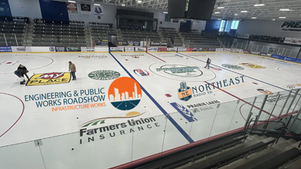Empty indoor ice rink with workers scraping the ice, large sponsor logos, bleachers, and a bright arena ceiling overhead.