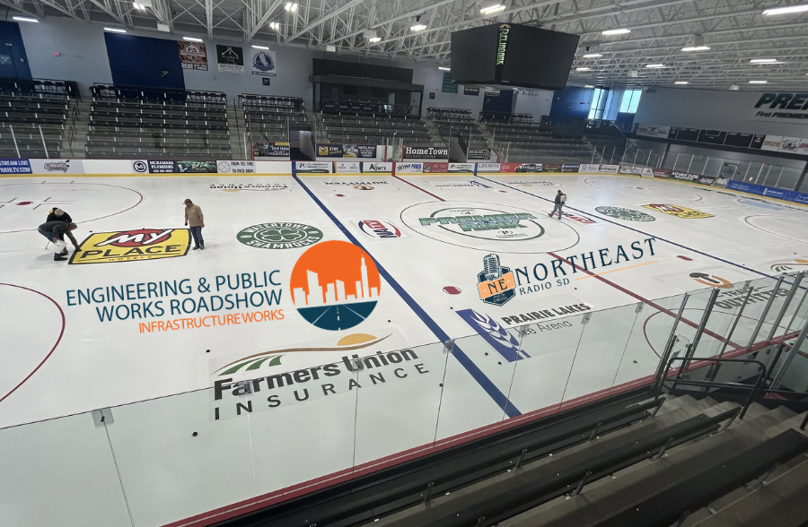 Empty indoor ice rink with workers scraping the ice, large sponsor logos, bleachers, and a bright arena ceiling overhead.