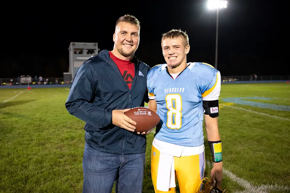 Two smiling men stand on a football field at night. One wears a blue and yellow jersey with number 8 and holds a football labeled "Player of the Game."