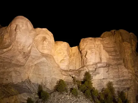 Lit Mount Rushmore at night against a dark sky. Four presidential faces carved in stone; trees below. Northeast Radio SD logo in corner.