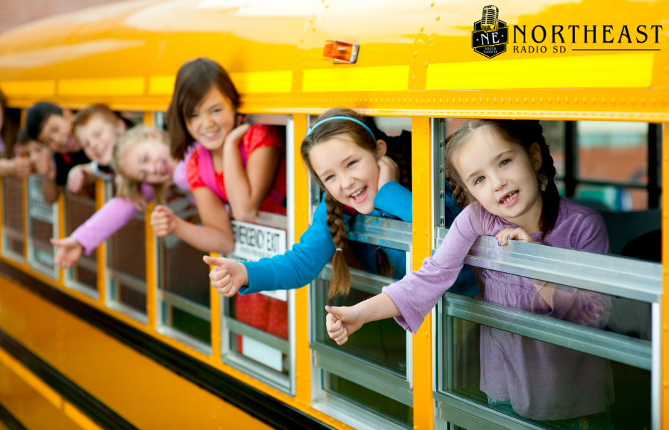 Smiling children lean from yellow school bus windows, giving thumbs up; NE Northeast Radio SD logo at top right.