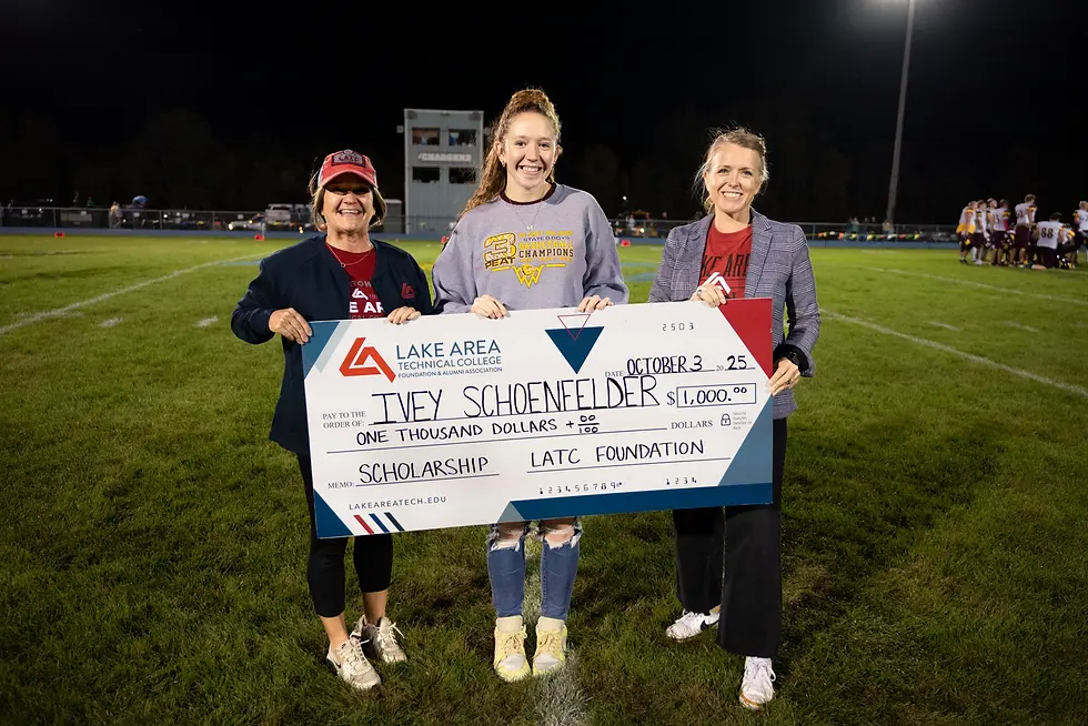 Three people holding a large $1,000 scholarship check on a football field at night. The mood is celebratory.