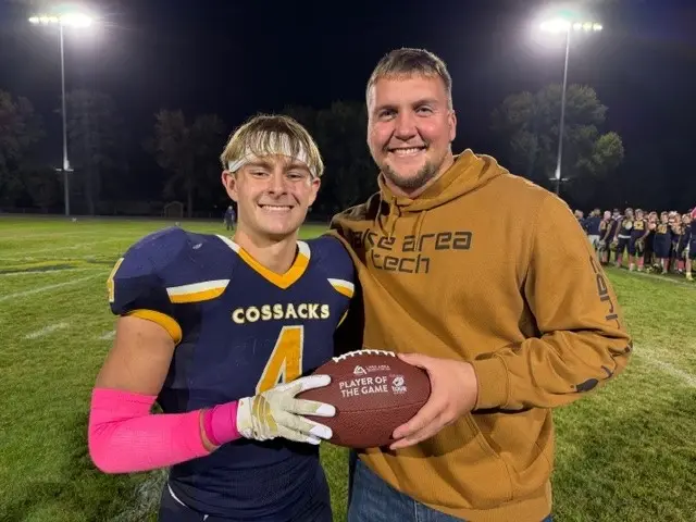 Smiling football player in blue and yellow uniform holds "Player of the Game" ball with a man in a brown hoodie on a lit field at night.