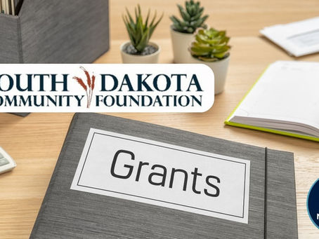 Office desk with a binder labeled "Grants," plants, calculator, and notebooks. Logo for South Dakota Community Foundation and Northeast Radio SD.
