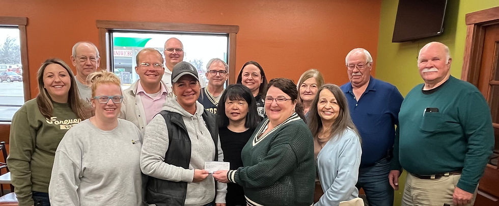 A group of 14 people smiling indoors. Two women in front are exchanging a check. The room has orange and green walls and windows.