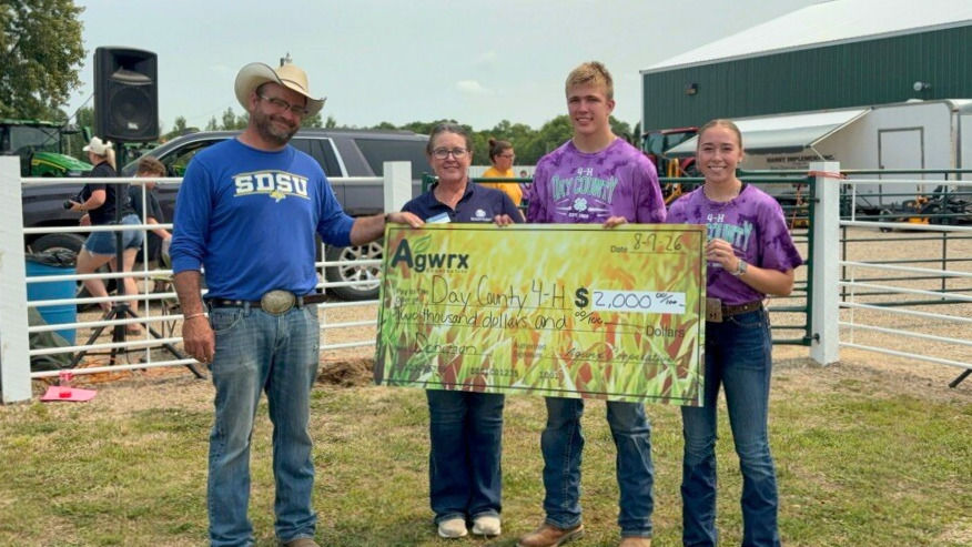 Four people hold a $2,000 Agwrx check for Day County 4-H at an outdoor fair, smiling in front of livestock pens.
