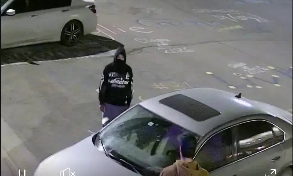 Person in black hoodie stands by a silver car at night. Chalk writings on the ground. Another person in a brown hoodie nearby.