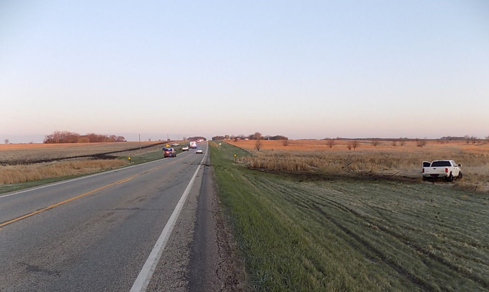 Rural road at sunset with a white truck off to the right in a field. Several cars, including one with flashing lights, are on the road.