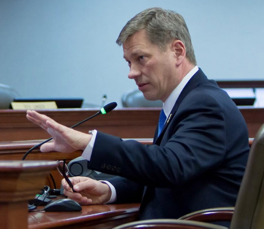 Man in a dark suit speaks at a microphone in a meeting room, gesturing with one hand and holding glasses, looking serious.