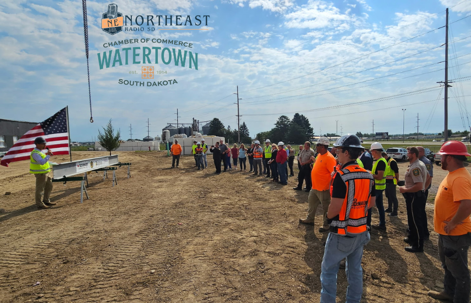 Workers and officials gather at a construction site as a speaker holds an American flag; Northeast Radio SD Watertown Chamber logo above.