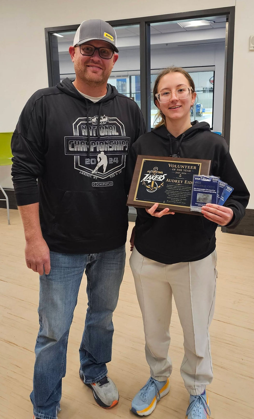 Man and young woman smile in a gym, holding a Volunteer of the Year plaque and Visa gift cards, both in black hoodies.