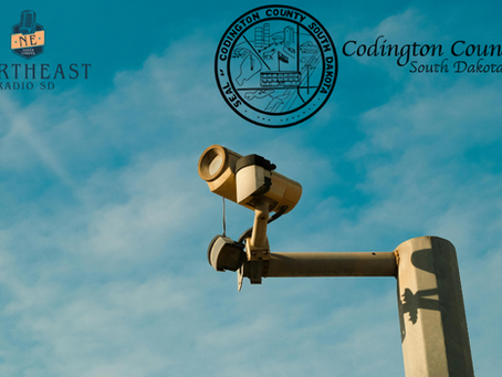Security camera on a pole against blue sky, with Northeast Radio SD and Codington County South Dakota logos and text.