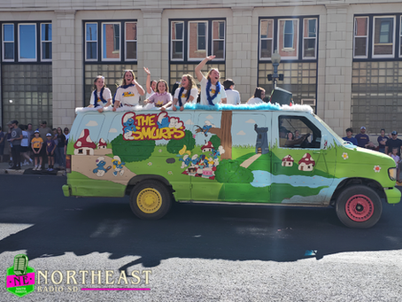 Girls wave from a Smurfs-themed parade van with colorful wheels on a sunny street, crowd behind, NE Northeast Radio SD logo visible.