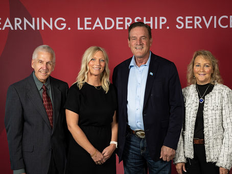 Four people smiling in front of a red backdrop with text: Learning. Leadership. Service. Business attire; friendly mood.