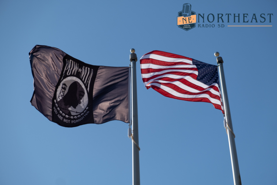 POW/MIA and U.S. flags wave on poles against a clear blue sky, with Northeast Radio SD logo at top right.