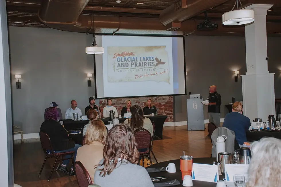 Panel discussion in a conference room. A screen displays "Glacial Lakes and Prairies." Attendees listen attentively. Cozy atmosphere.