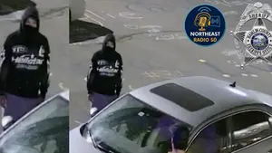 Person in a black hoodie and mask stands near a parked gray car on a marked concrete surface. Sheriff's badge and Northeast Radio SD logos are visible.
