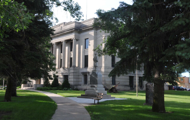 Stone civic building with columns behind trees, a statue on a lawn, and a curved sidewalk in a quiet park-like setting.