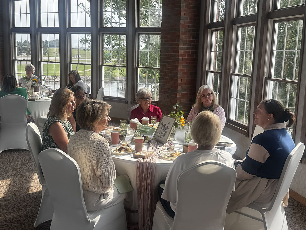 Eight women chat around a tea table in a sunlit windowed room, with flowers, pastries, and a calm, friendly mood.